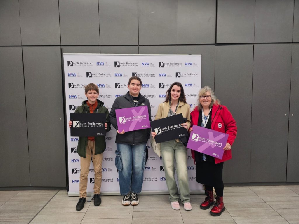 Somerset Members of Youth Parliament James Lawson, Emma Brown and Ellie Bealing, standing in a line with Somerset Council Partnerships Manager Fiona Phur. All are holding signs with 'Youth Parliament: Making Our Mark' written on them.