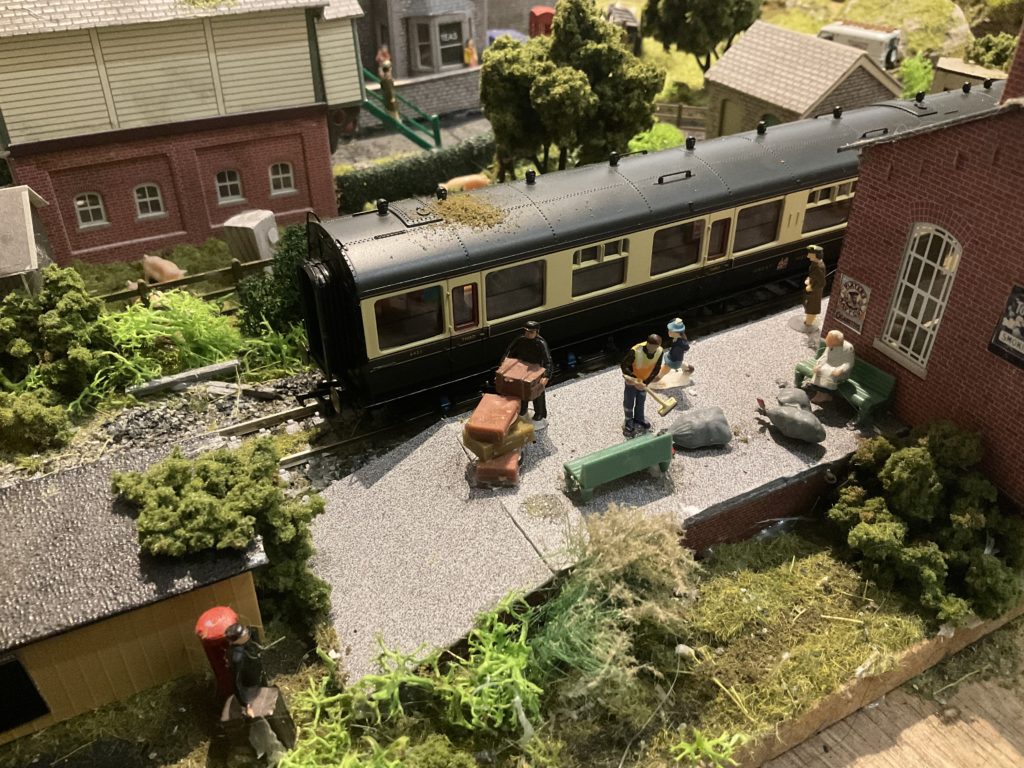 A close up of a model train and station, with miniature porter loading luggage, cleaner sweeping the platform, child running, woman waiting for the train, older lady feeding birds.